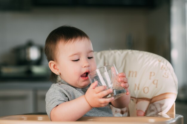 Nettes kleines Mädchen, das in einem Babystuhl in der Küche und im Trinkwasser sitzt