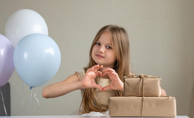 Nettes kleines Mädchen, das mit ihren Händen Herz macht und lächelt, umgeben von Luftballons und Geschenken