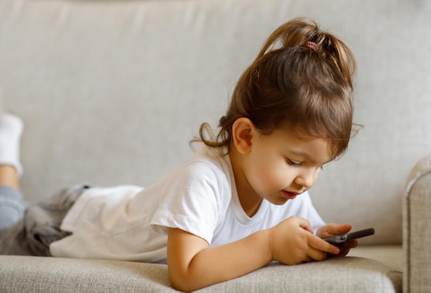 Nettes kleines Mädchen, das Online-Spiele auf dem Smartphone spielt, während es auf der Couch liegt
