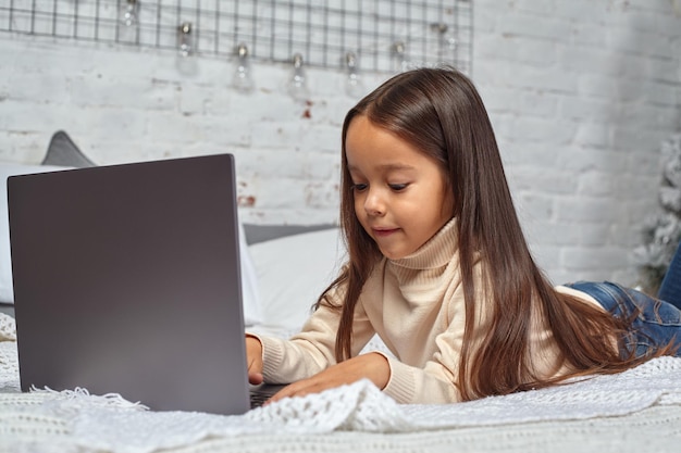 Nettes kleines Mädchen, das sich amüsant fühlt, während es Cartoons auf einem Laptop sieht, der auf dem Bett sitzt