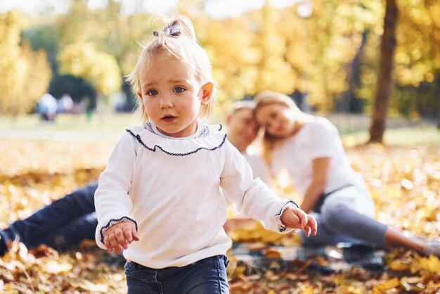 Nettes kleines Mädchen geht zur Kamera. Fröhliche junge Familie erholen sich zusammen in einem Herbstpark.