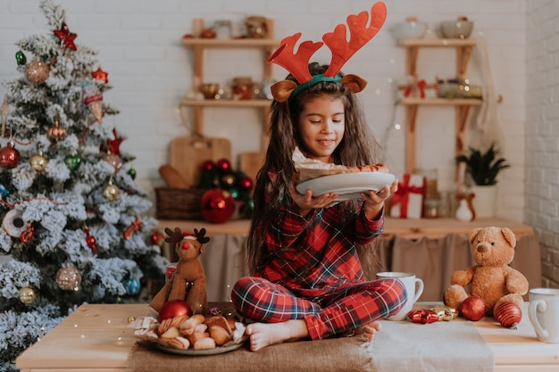 Nettes kleines Mädchen im karierten roten Pyjama mit Hirschhörnern auf dem Kopf isst einen Weihnachtskuchen