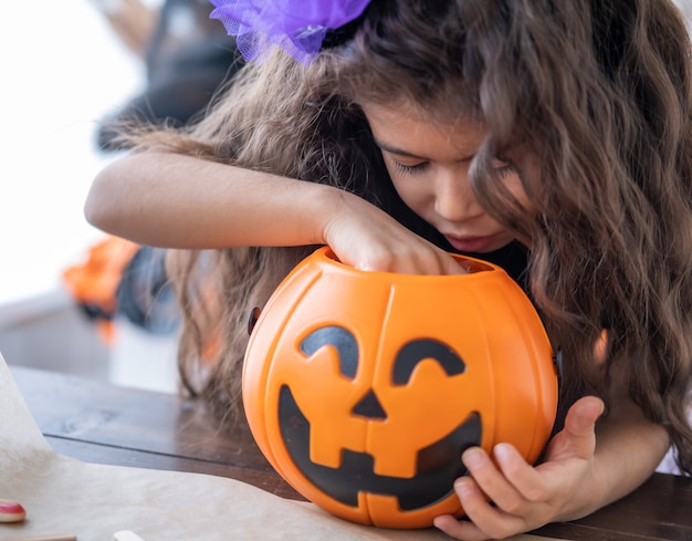 Nettes kleines Mädchen im Kostüm der Hexe, die Kürbissteckfassung mit Bonbons hält, Spaß in der Küche hat und Halloween feiert.