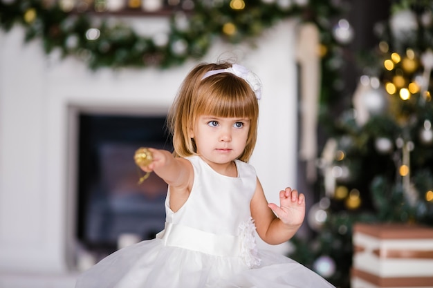 Nettes kleines mädchen im weißen kleid mit nettem kranz nahe dem weihnachtsbaum