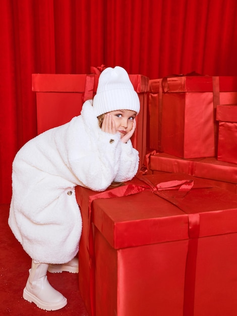 Nettes kleines Mädchen im weißen Mantel neben den riesigen roten Weihnachtsgeschenkboxen auf rotem Hintergrund. Feiertage