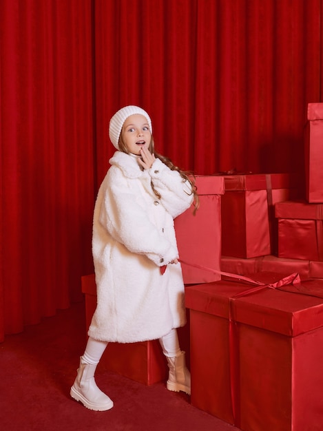 Nettes kleines Mädchen im weißen Mantel und hatte die riesigen roten Weihnachtsgeschenkboxen auf rotem Hintergrund