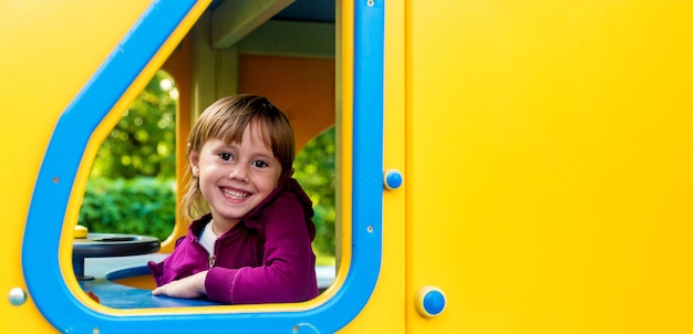 Nettes kleines Mädchen in den dunkelrosa Kleidern, die vom Spielplatzspielzeug am Park herausschauen