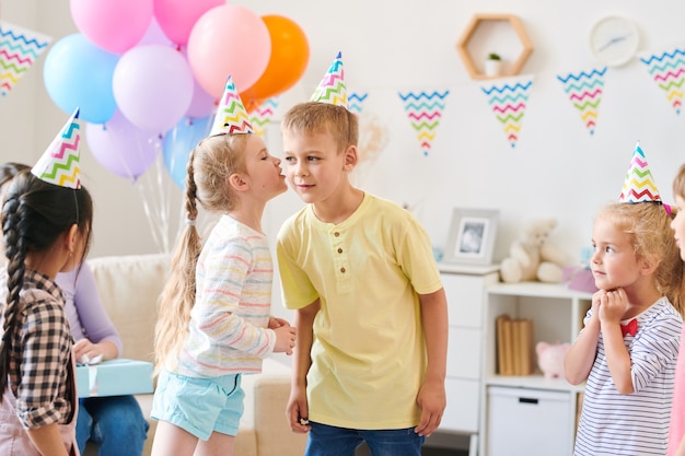 Nettes kleines Mädchen in der Freizeitkleidung, das etwas zum Jungen flüstert, während Spiel mit Freunden zu Hause Geburtstagsfeier spielt