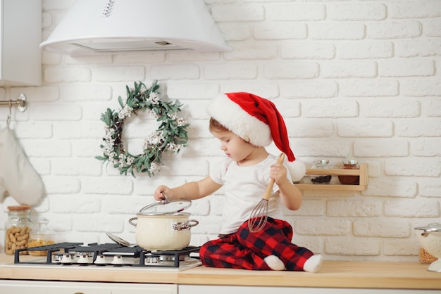 Nettes kleines Mädchen in der Weihnachtsmütze, das Kekse in der Küche zu Hause vorbereitet. Sitzt auf dem Küchentisch und hilft Mama, ein festliches Weihnachtsessen vorzubereiten