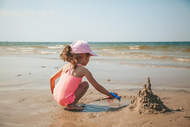 Nettes kleines Mädchen in einem rosafarbenen Badeanzug, der mit Sand am Strand einer Ostsee spielt