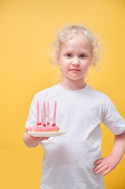 Nettes kleines Mädchen in einem weißen T-Shirt, das einen Donut mit Kerzen hält, Spaß hat und ein Kind in einem Strohhut Gesichter macht