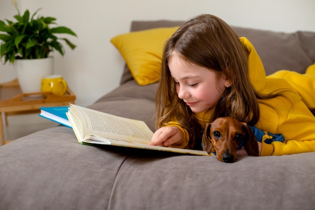 Nettes kleines Mädchen in gelber Kleidung liest ein Buch, das mit einem Zwergdackel auf dem Bett liegt