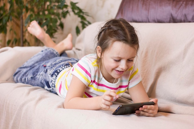 Nettes kleines Mädchen liegt zu Hause auf dem Sofa, lächelt und benutzt ein Smartphone