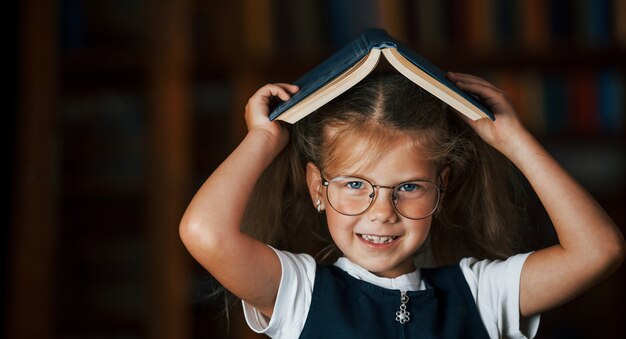 Nettes kleines Mädchen mit Brille steht in der Bibliothek voller Bücher