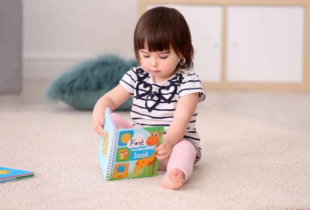 Nettes kleines Mädchen mit Buch, das zu Hause auf dem Boden sitzt