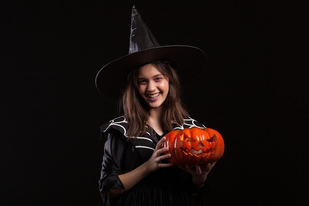Nettes kleines Mädchen mit einem bösen Lachen, das Zauberei mit einem Kürbis für Halloween tut. Entzückendes Kind in einem Hexenkostüm für Halloween.