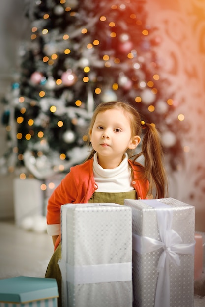Nettes kleines Mädchen mit einem Weihnachtsgeschenk an einem Weihnachtsbaum im gemütlichen Wohnzimmer