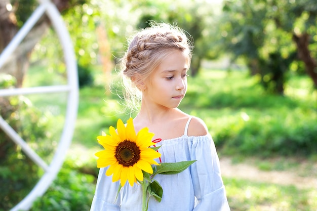 Nettes kleines Mädchen mit einem Zopf auf dem Kopf hält eine Sonnenblume im Sommergarten.