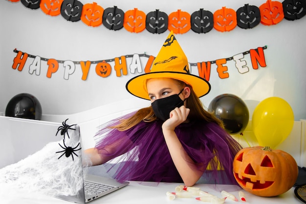 Nettes kleines Mädchen mit Hexenhut, das hinter dem Tisch in einem mit Halloween-Thema dekorierten Raum sitzt Covid19