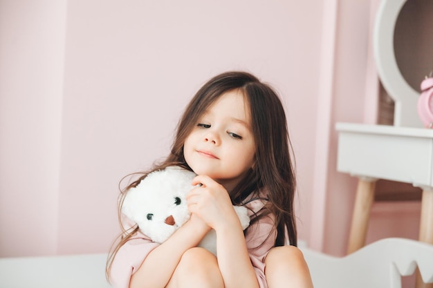 Nettes kleines Mädchen mit langen Haaren umarmt einen Teddybären, schaut auf die Kamera und lächelt, während sie zu Hause auf ihrem Bett sitzt