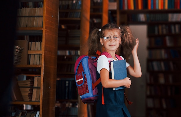 Nettes kleines Mädchen mit Rucksack steht in der Bibliothek voller Bücher