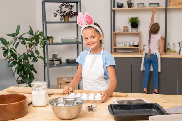 Nettes kleines Mädchen mit Schürze, das reife Tomaten an die ältere Schwester weitergibt, während sie gemeinsam köstlichen Salat kocht, Kücheneinrichtung im Hintergrund