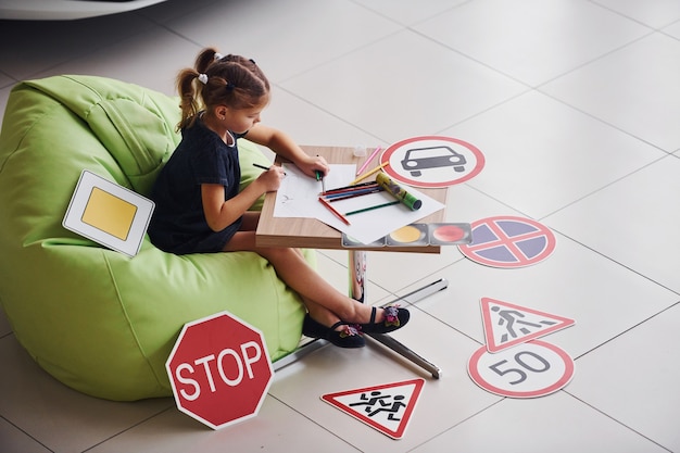 Nettes kleines Mädchen sitzt auf dem weichen grünen Stuhl am Tisch mit Bleistift- und Papierblättern. In der Nähe von modernen Auto- und Verkehrsschildern auf dem Boden.