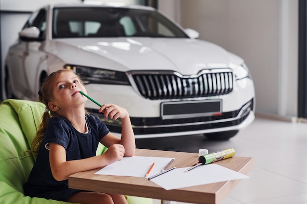 Nettes kleines Mädchen sitzt auf dem weichen grünen Stuhl am Tisch mit Bleistift- und Papierblättern. Nahe bei modernen Automobilen.