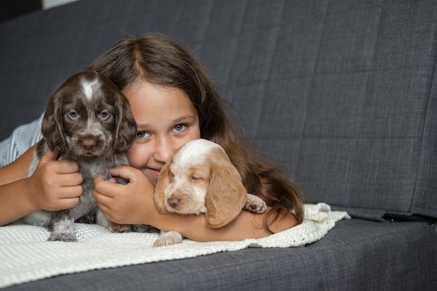 Nettes kleines Mädchen umarmt und liegt mit zwei russischen Spaniel-Hündchen unter auf der Couch. Haustierbetreuung und freundliches Konzept. Liebe und Freundschaft zwischen Mensch und Tier.