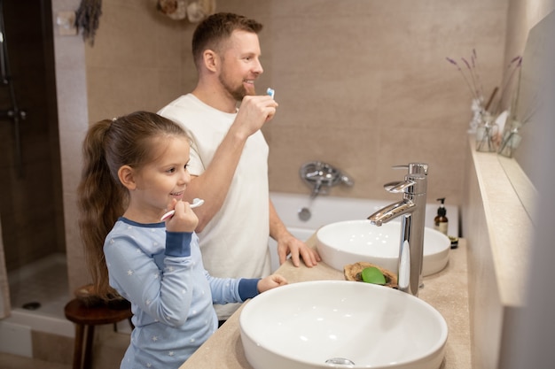 Nettes kleines Mädchen und ihr glücklicher junger Vater mit Zahnbürsten, die Zähne putzen, während sie im Badezimmer vor dem Spiegel stehen