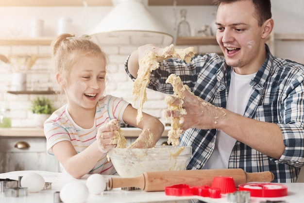 Foto nettes kleines mädchen und ihr vater kneten teig und lächeln beim backen in der küche zu hause. vatertag, familie, urlaubskonzept