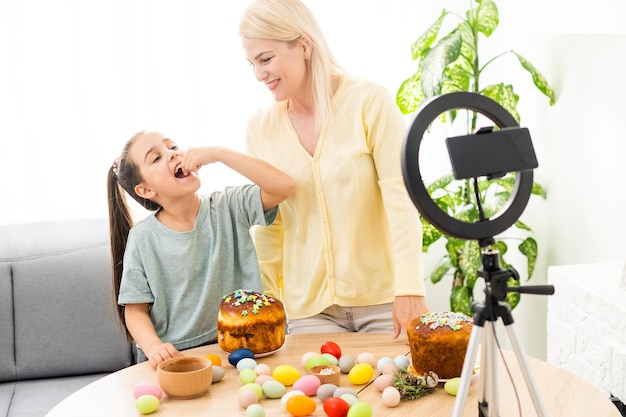 Nettes kleines Mädchen und schöne junge Mutter sitzen zusammen am Küchentisch mit Smartphone für online zu Ostern.