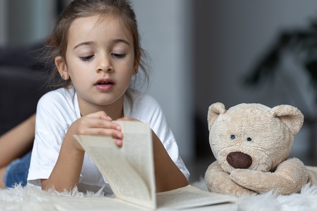 Nettes kleines Mädchen zu Hause, das mit ihrem Lieblingsspielzeug auf dem Boden liegt und ein Buch liest.