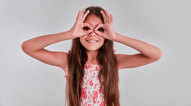 Foto nettes kleines mädchenporträt auf grauem hintergrund, faltete ihre finger wie eine brille über ihren augen und lächelte.