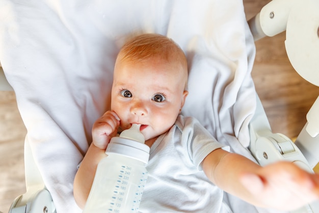 Nettes kleines neugeborenes mädchen trinkt milch aus der flasche und schaut in die kamera auf weißem hintergrund...