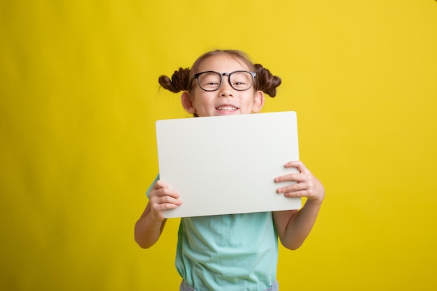 Nettes kleines Schulmädchen, das ein weißes Blatt Papier auf gelbem Hintergrund hält