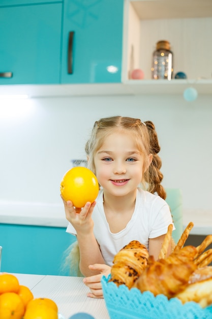 Nettes kleines verspieltes lustiges Mädchen, das glücklich orange Orangen hält, mit Küche im Hintergrund