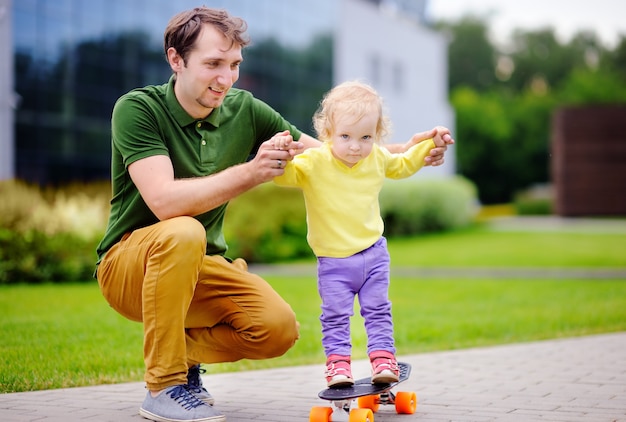 Nettes Kleinkindmädchen, das lernt, mit ihrem Vater draußen Skateboard zu fahren