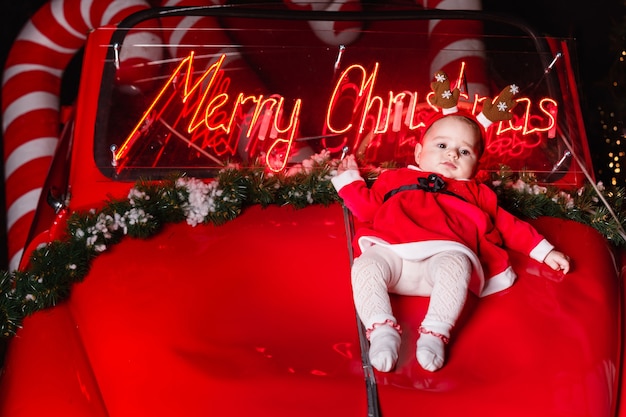 Nettes Kleinkindmädchen mit im Hirschgeweih, das auf Retro rotem Auto am Weihnachtsstudio liegt. Speicherplatz kopieren. heiraten Weihnachten auf Hintergrund.