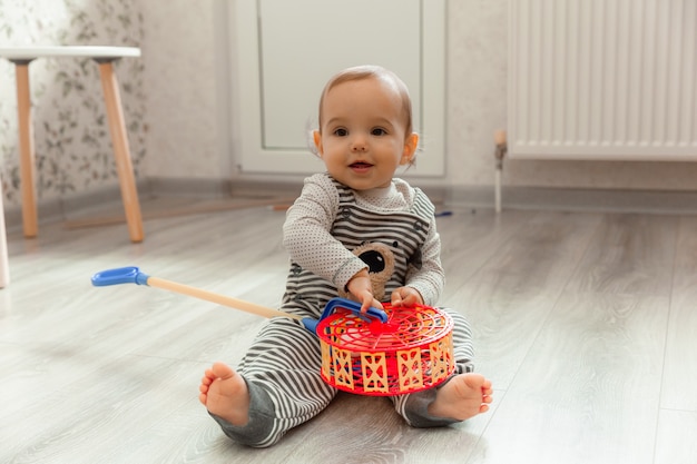 Nettes lächelndes Baby 12 Monate alt sitzt auf dem Boden in seinem Zimmer und spielt mit Spielzeug.