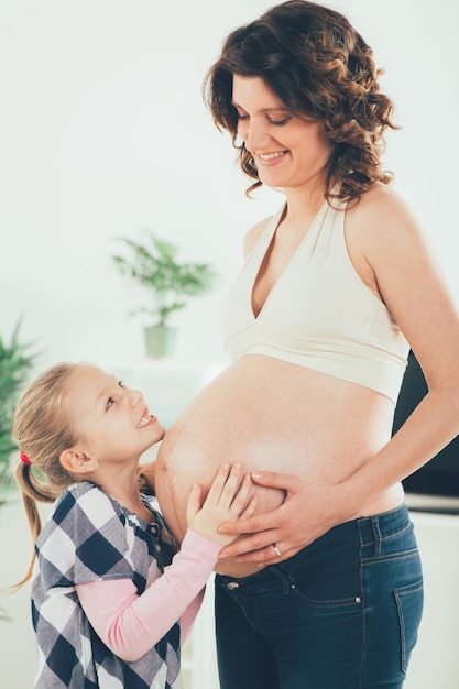 Nettes lächelndes junges Mädchen, das den schwangeren Bauch ihrer schwangeren Mutter kuschelt.