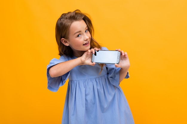 Nettes lächelndes junges Mädchen in einem blauen Kleid zeigt das Telefon in horizontaler Position mit einem Modell auf einem gelben Hintergrund