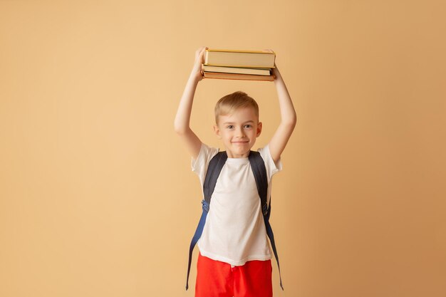 Nettes lächelndes Kind mit einem Rucksack, der Bücher zur Vorbereitung auf die Schule auf beigem Hintergrund hält