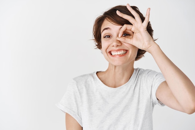 Nettes lächelndes Mädchen, das okayzeichen auf Auge zeigt und glücklich schaut, das perfekte Produkt lobend, das im T-Shirt gegen weißen Hintergrund steht