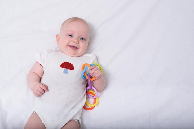 Nettes lächelndes neugeborenes Baby, das auf weißen Laken mit Spielzeug in der Hand liegt Draufsicht
