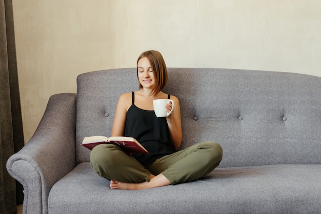 Nettes liebliches lächelndes Mädchen, das ein Buch liest und Tee trinkt, während auf dem Sofa zu Hause entspannt. Hobby Lebensstil.