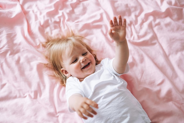 Nettes lustiges Babymädchen in weißer Kleidung auf dem Bett zu Hause, Blick von oben