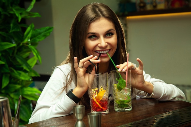 Foto nettes lustiges brünettes mädchen trinkt zwei cocktails gleichzeitig und lächelt vorne