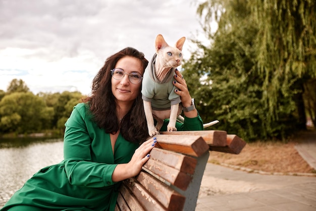 Nettes Mädchen, das eine pelzige Katze im Freien streichelt Sitzt auf einer Holzbank mit grünen Bäumen und Flusshintergrund Liebe zu den Tieren