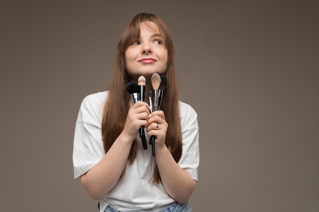 Nettes Mädchen hält Make-up-Pinsel nahe dem Kinn an einer dunklen Wand